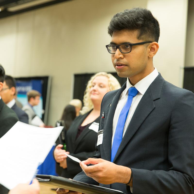 A student wearing glasses and a suit holding a padfolio speaking with a recruiter at a Drake University career fair
