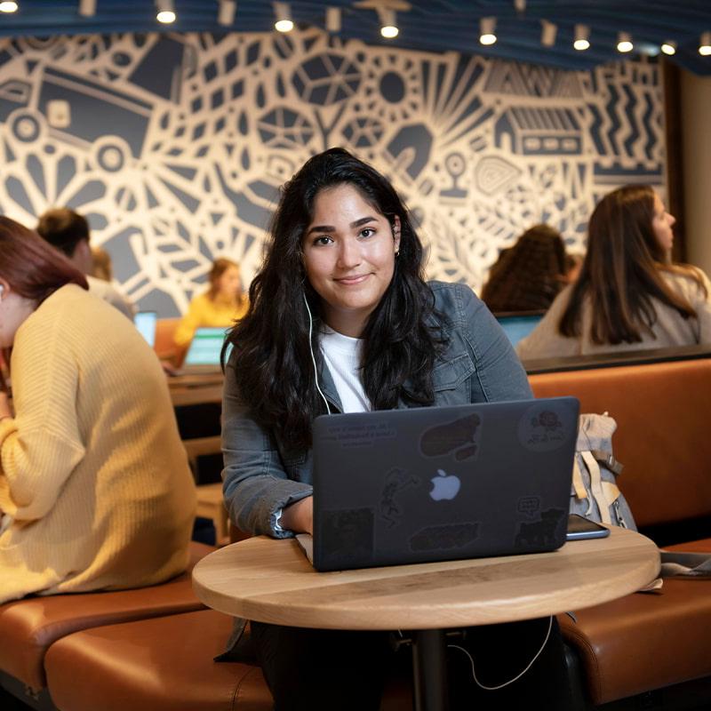 威尼斯人网上娱乐的一名学生坐在校园餐厅的一张桌子上，一边用笔记本电脑工作，一边微笑着